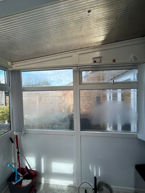 Interior view of a sunlit enclosed porch with large glass doors, white walls, corrugated metal ceiling, and brick exterior visible through windows
