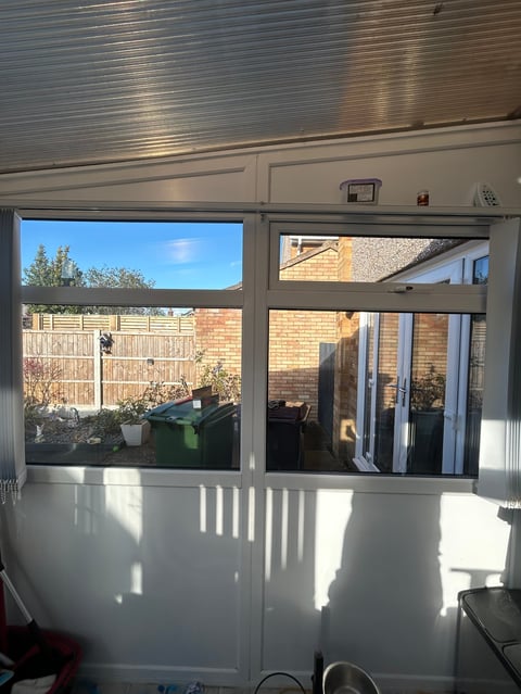Sunlit room with large sliding glass doors and windows overlooking a backyard with wooden fence and green trash bins