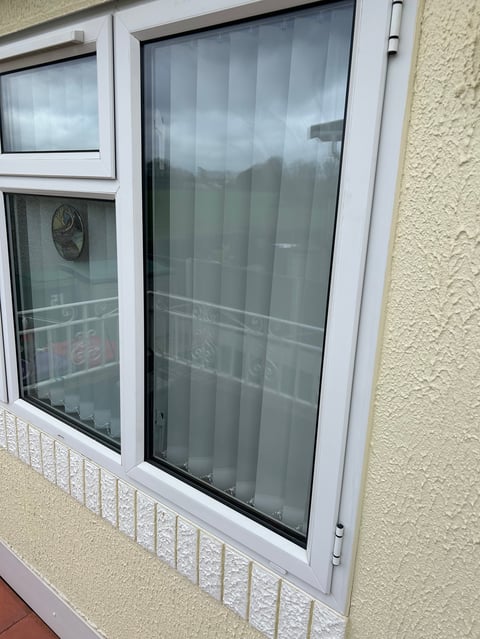 Two white-framed windows with vertical blinds on a beige textured wall, with white metal grating below